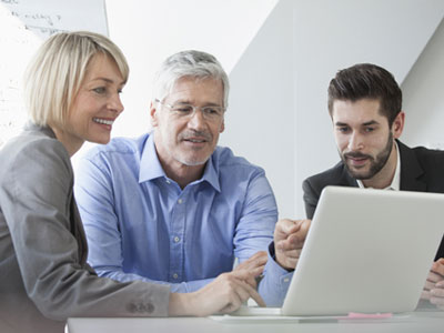 Three adults sitting at table, looking at laptop screen.