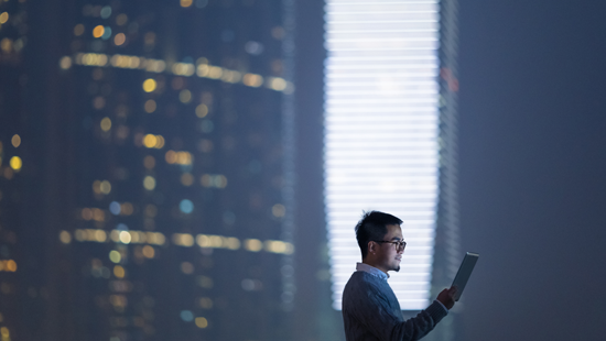 Man looking at tablet with city skyline at night in the background.