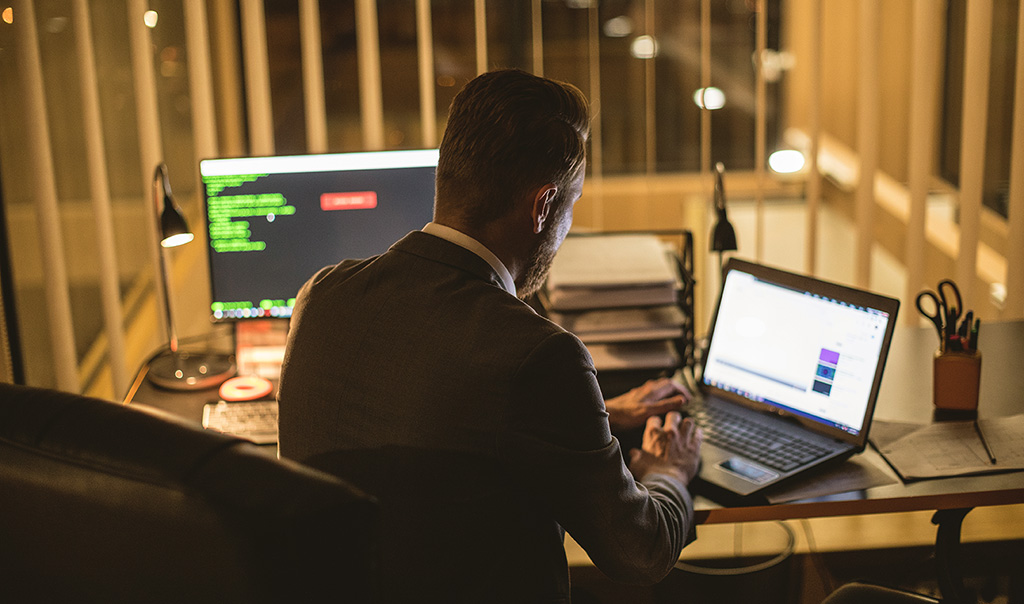 Image of man sitting at a desk in front of a desktop computer while working on his laptop to the side