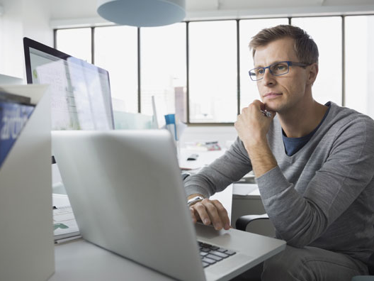 Man working on laptop computer in bright office with many windows.