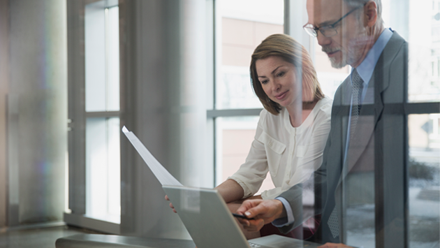 Woman and man conferring over laptop at table in glass-walled office. 