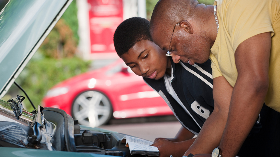 A Dad and his teenage son leaning over a car engine, reading a car maintenance manual.