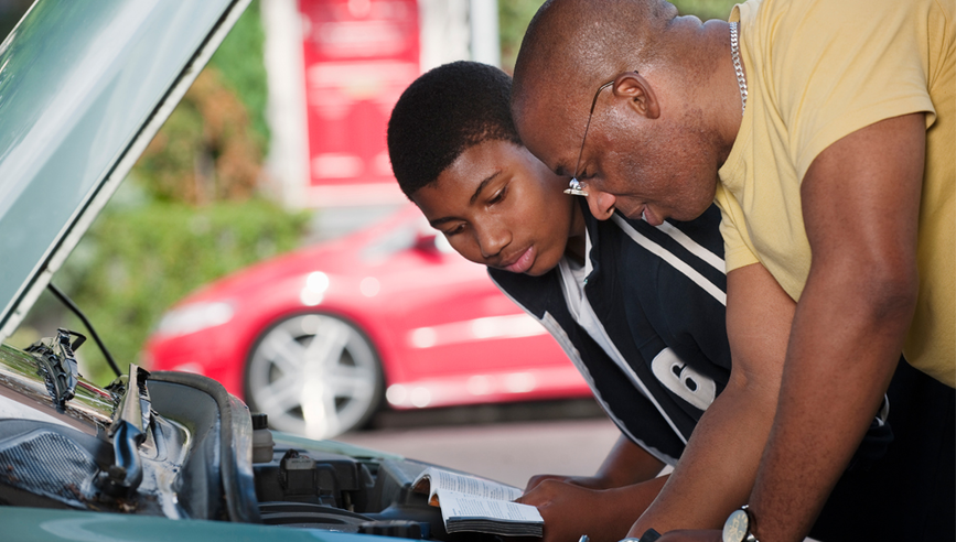 A Dad and his teenage son leaning over a car engine, reading a car maintenance manual.