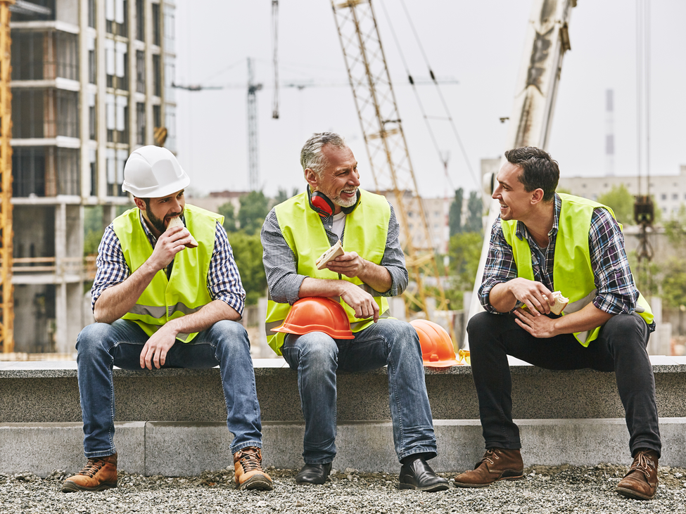 Group of builders in working uniform are eating sandwiches and talking while sitting on stone surface against construction site