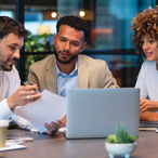 Three business people meeting and looking at a laptop and a document. There is paperwork and other technology on the table, formal business wear.