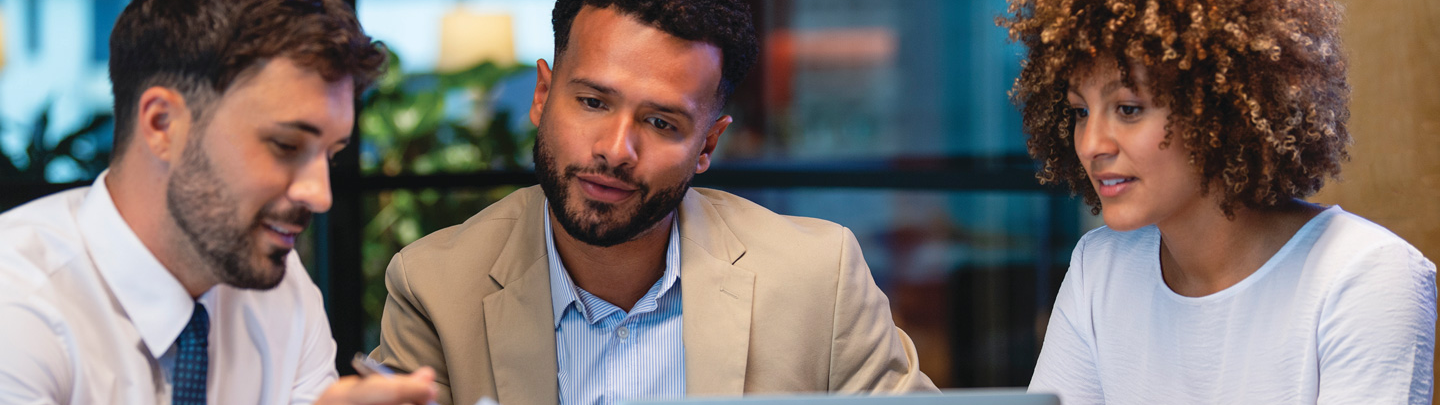 Three business people meeting and looking at a laptop and a document. There is paperwork and other technology on the table, formal business wear.