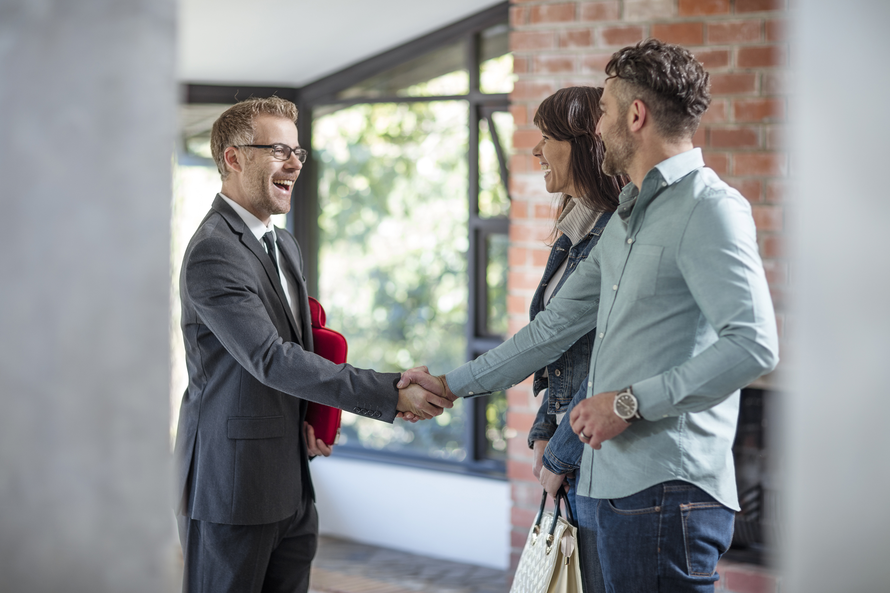Man in suit shaking hands with couple wearing casual clothing in office building hallway.