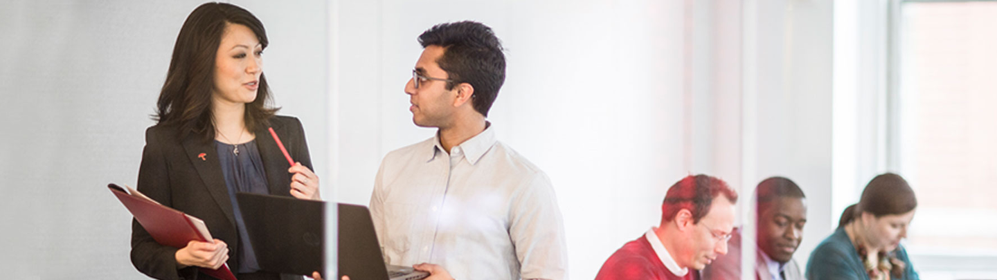 Woman and man talking at the front of a conference room, people sitting at table in background.