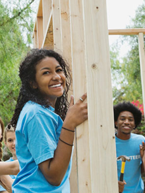 Smiling young women volunteers in matching sky-blue shirts raising framing on home build site.