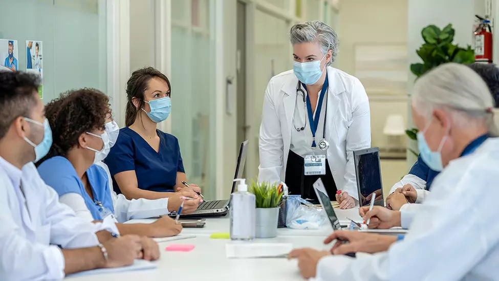 A medical professional standing in front of a table seated by other people in masks.