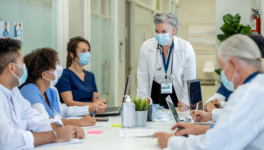 A medical professional standing in front of a table seated by other people in masks.