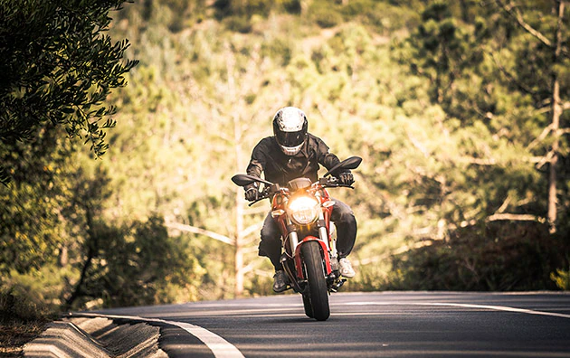 A lone motorcyclist driving down a country road on a sunny day, motorcycle operator wearing a helmet, leather jacket and gloves.