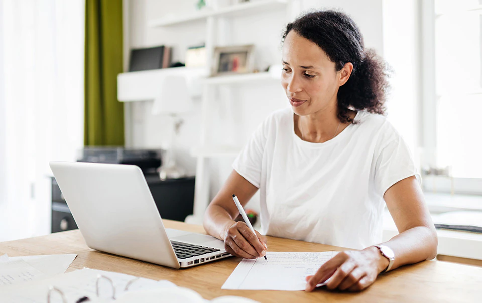 A woman sitting at a table reviewing car insurance quotes on laptop and taking notes.