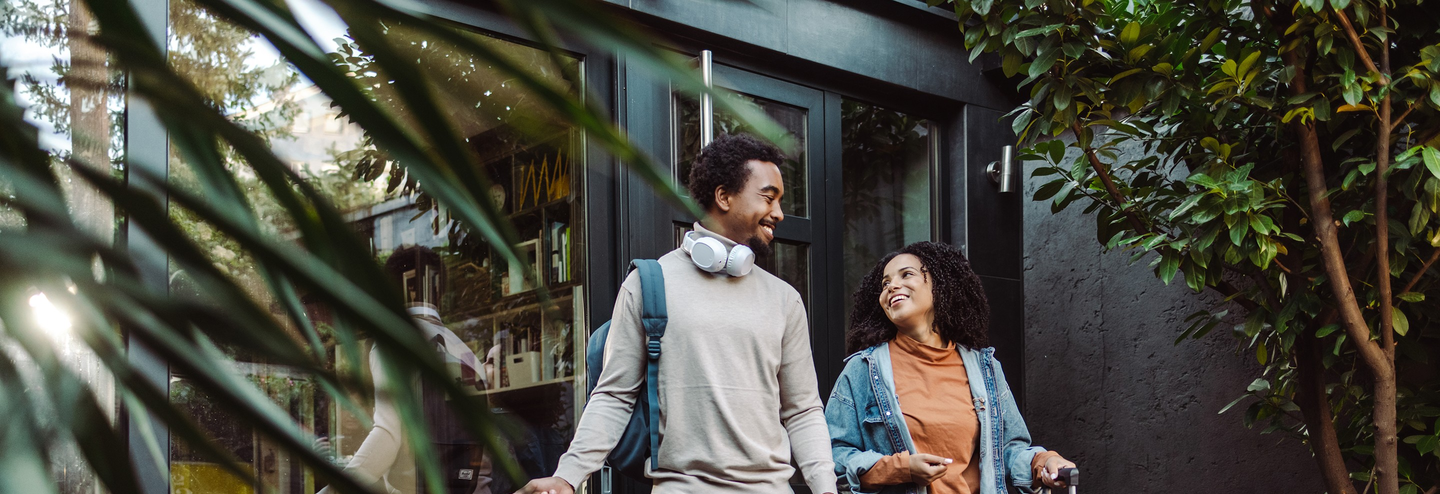 An excited couple holding luggage smile as they step out of a house.