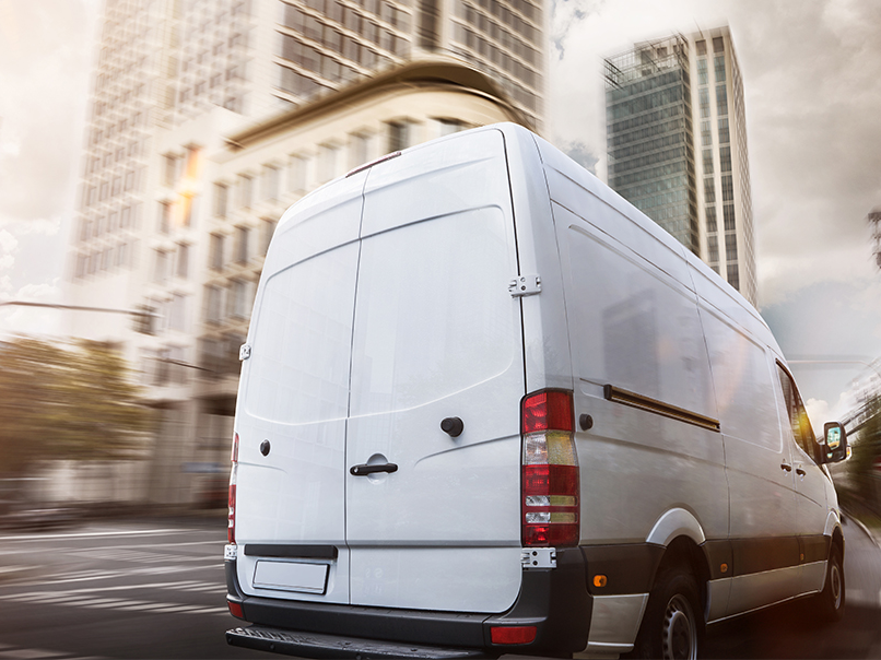 A white business delivery van driving on a downtown city street.