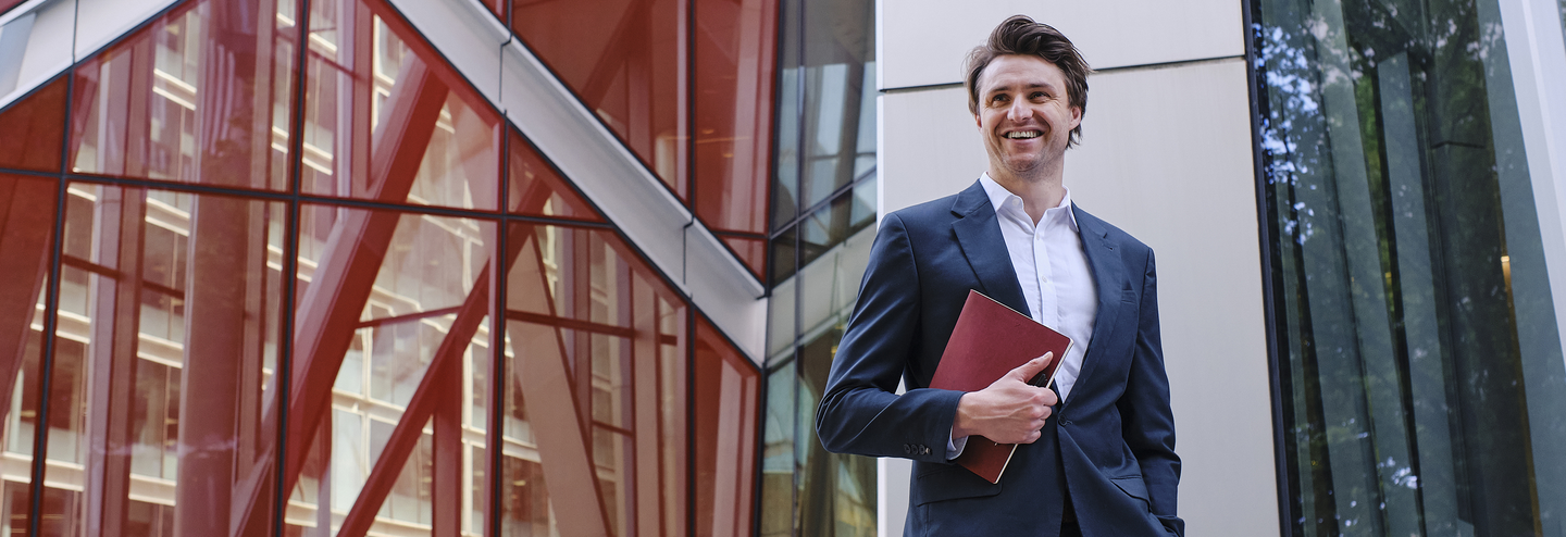 A smiling businessman stands holding his diary in front of a contemporary office building.