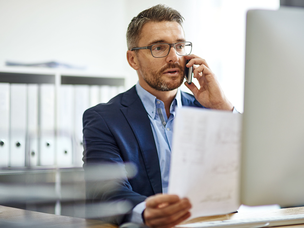 Businessman reads document while working on phone and computer.