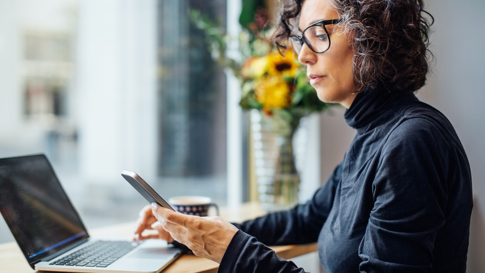 Mature businesswoman sitting at cafe looking at her cell phone while working on laptop computer.