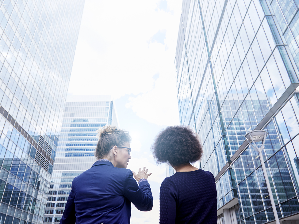 Two businesspeople stand outside in a modern office plaza as one gestures toward a corridor of skyscrapers whilst discussing a concept with her colleague