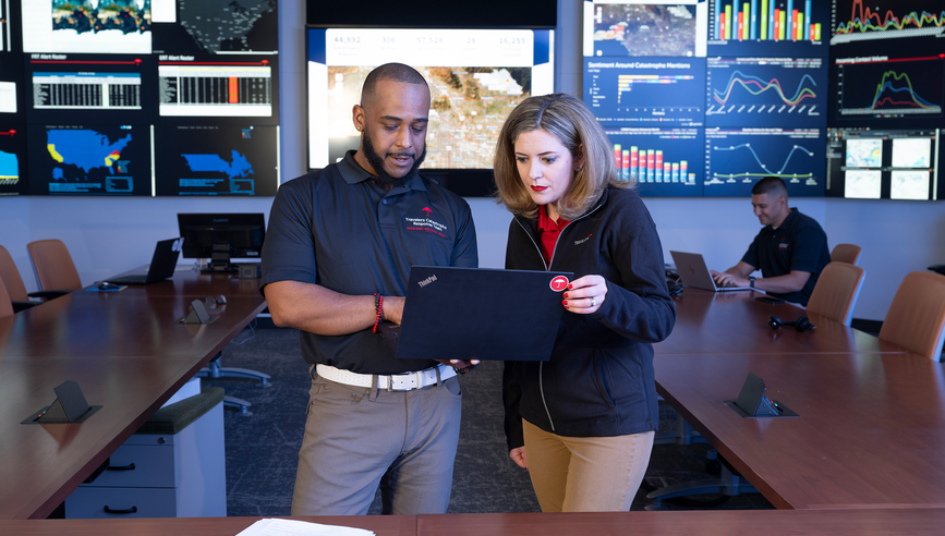 Man and woman looking at a laptop with data on monitors behind them.