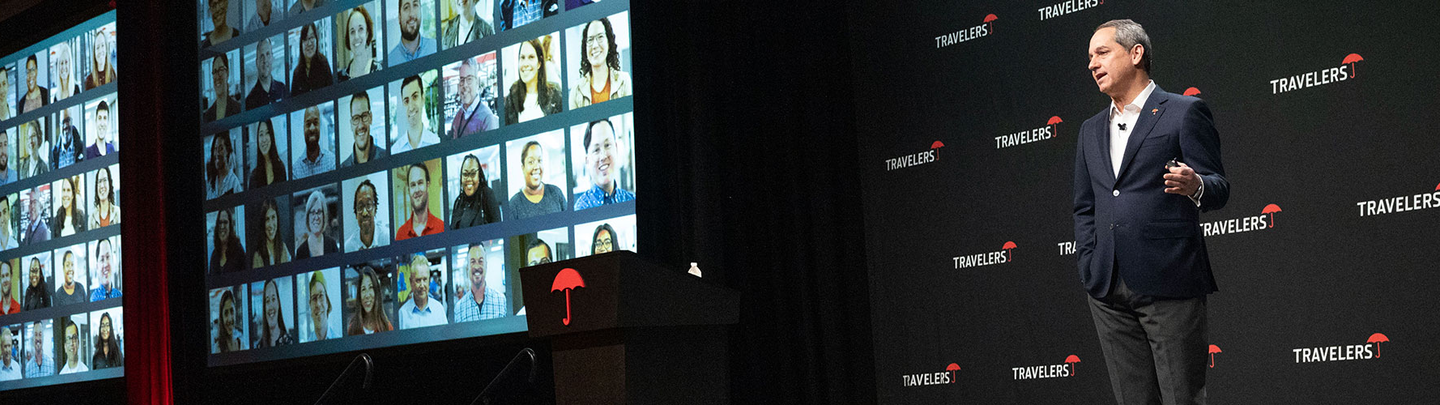 Travelers CEO, Alan Schnitzer, speaking on stage in front of a screen with headshots of Travelers employees.