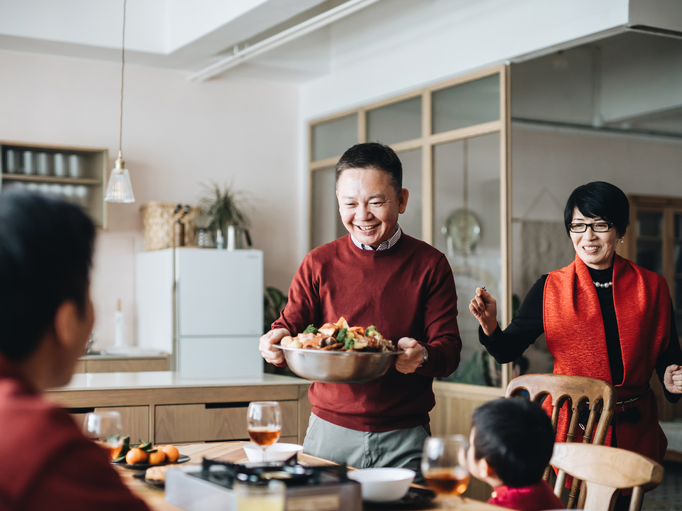 A man joyfully brings a tasty stir fry dish to his family seated around a kitchen table.