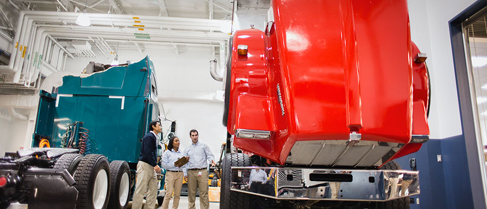 Claim department female with a clipboard talking with two male coworkers. They are standing between a green truck and a red truck in a large warehouse.