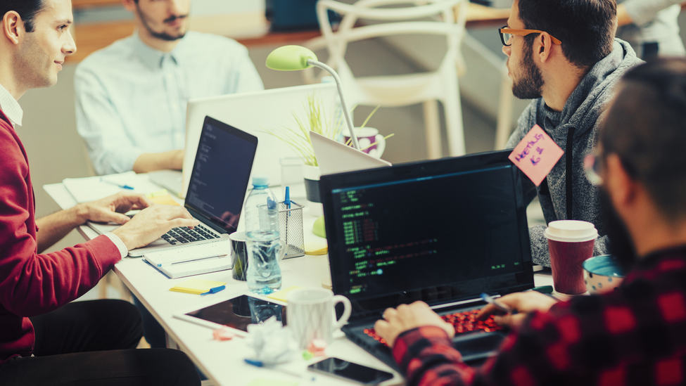 Five young people running their coding startup company. Working from the house like business incubator. Sitting and collaborating. Using their laptops for coding. Selective focus to young man with glasses talking to his coworker.