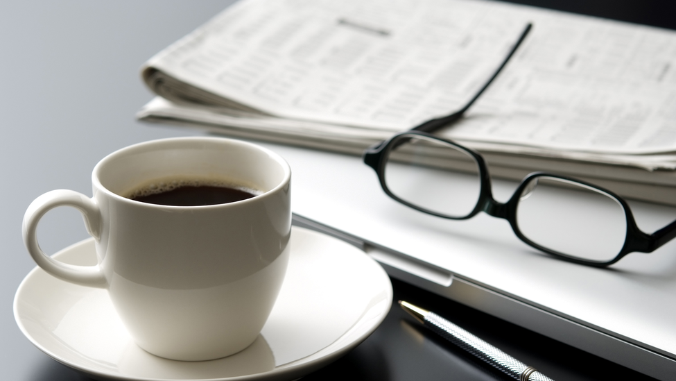 A close up of a desk at a business office. This desk includes a cup of black coffee served in a white cup on a white saucer, a laptop computer, a pair of glasses and newspaper.