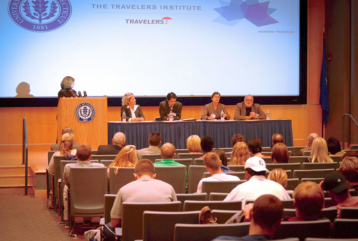 Attendees in the audience watch one speaker at a podium and four seated panelists at the "Overdraft" Symposia Series event at the University of Connecticut