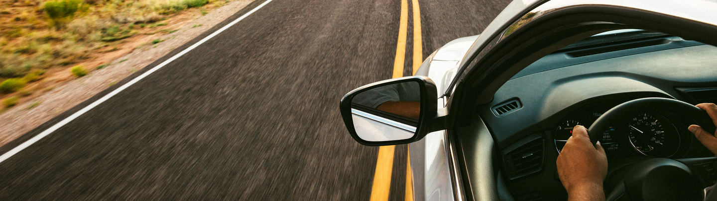 person inside a car, both hands on the steering wheel, driving on an open road