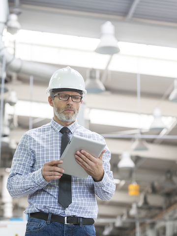 A health and safety inspector wearing a hard hat carries a clipboard while inspecting an industrial building.