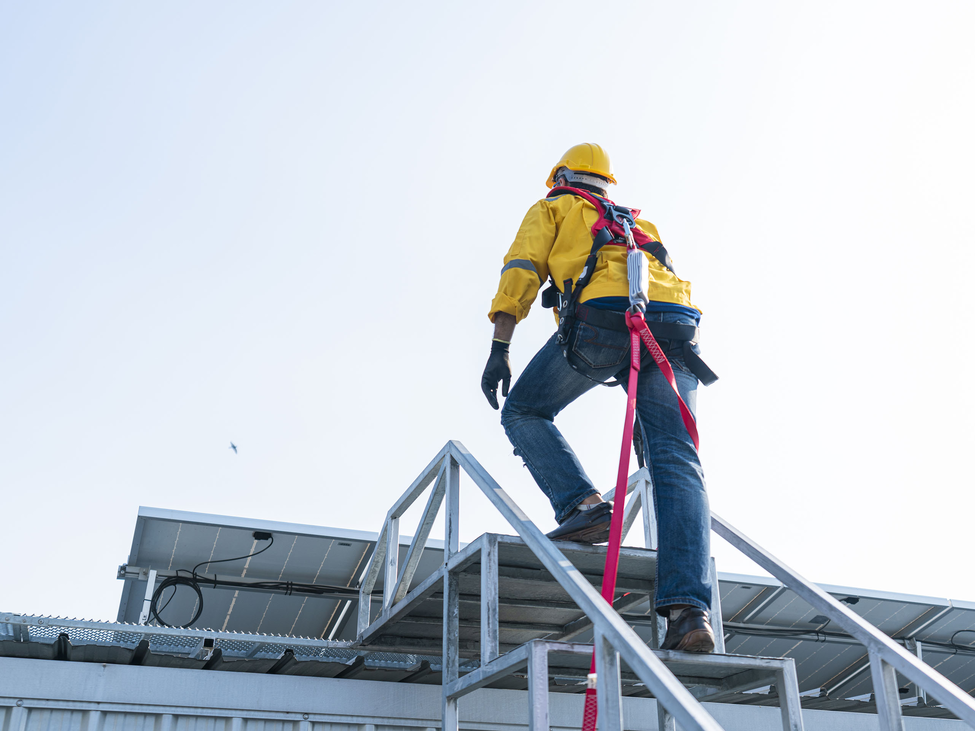 Construction worker is tied in as he climbs metal steps on a roof with solar panels.