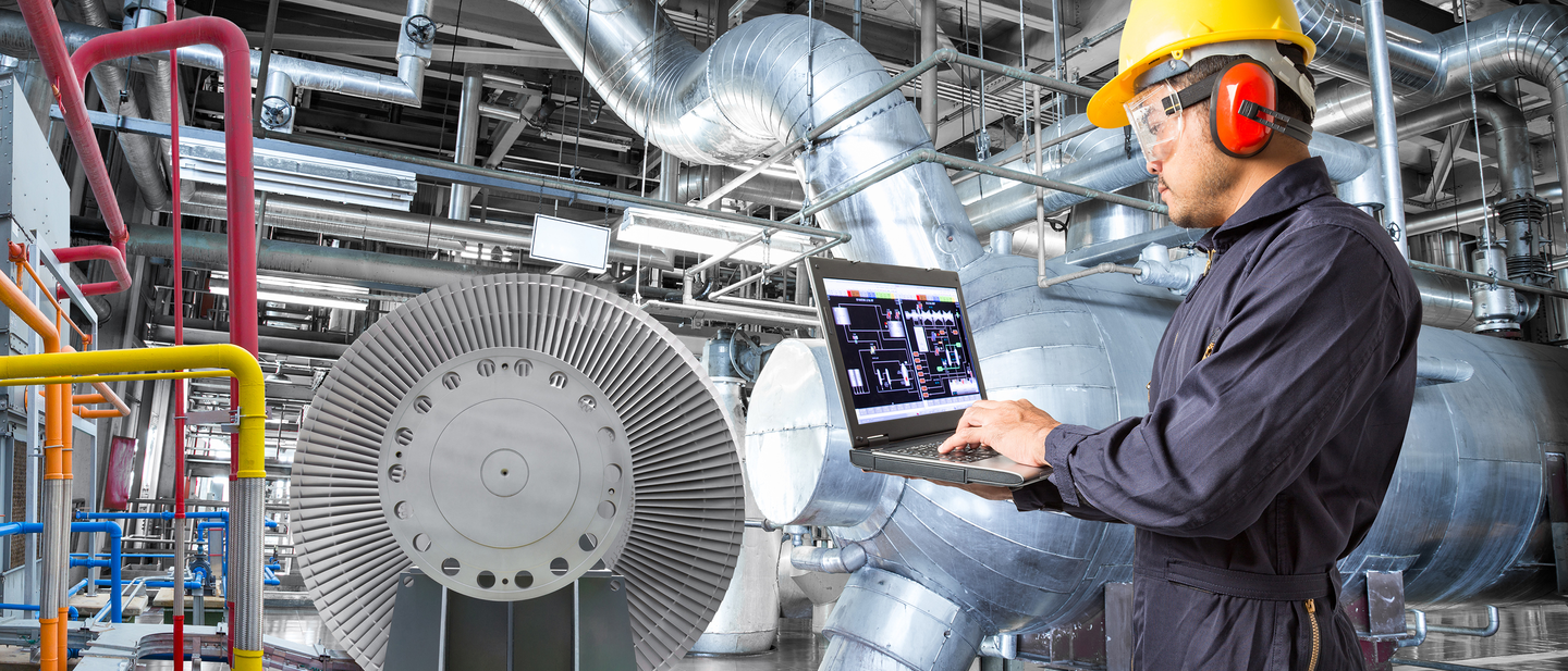 A man wearing eye and ear safety equipment looking at a laptop screen in a manufacturing plant.