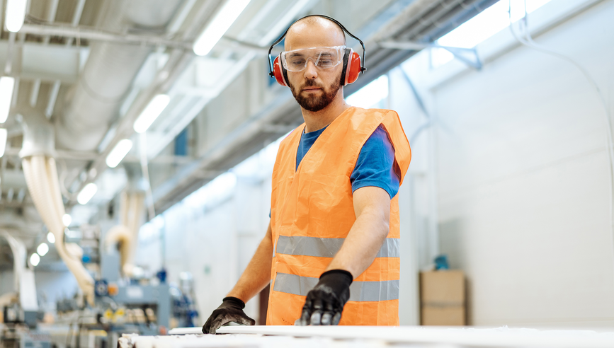 factory worker wearing orange vest, safety goggles, ear muffs and working at a conveyor belt