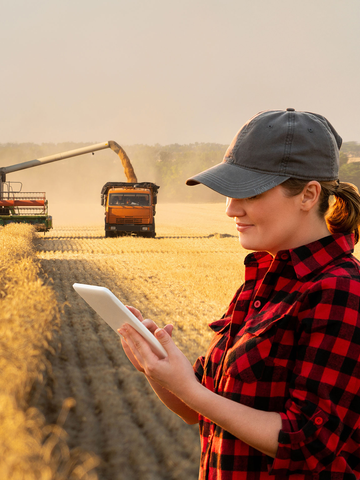 Farmer in buffalo-check shirt holding up tablet computer, standing in field with harvester in the background.