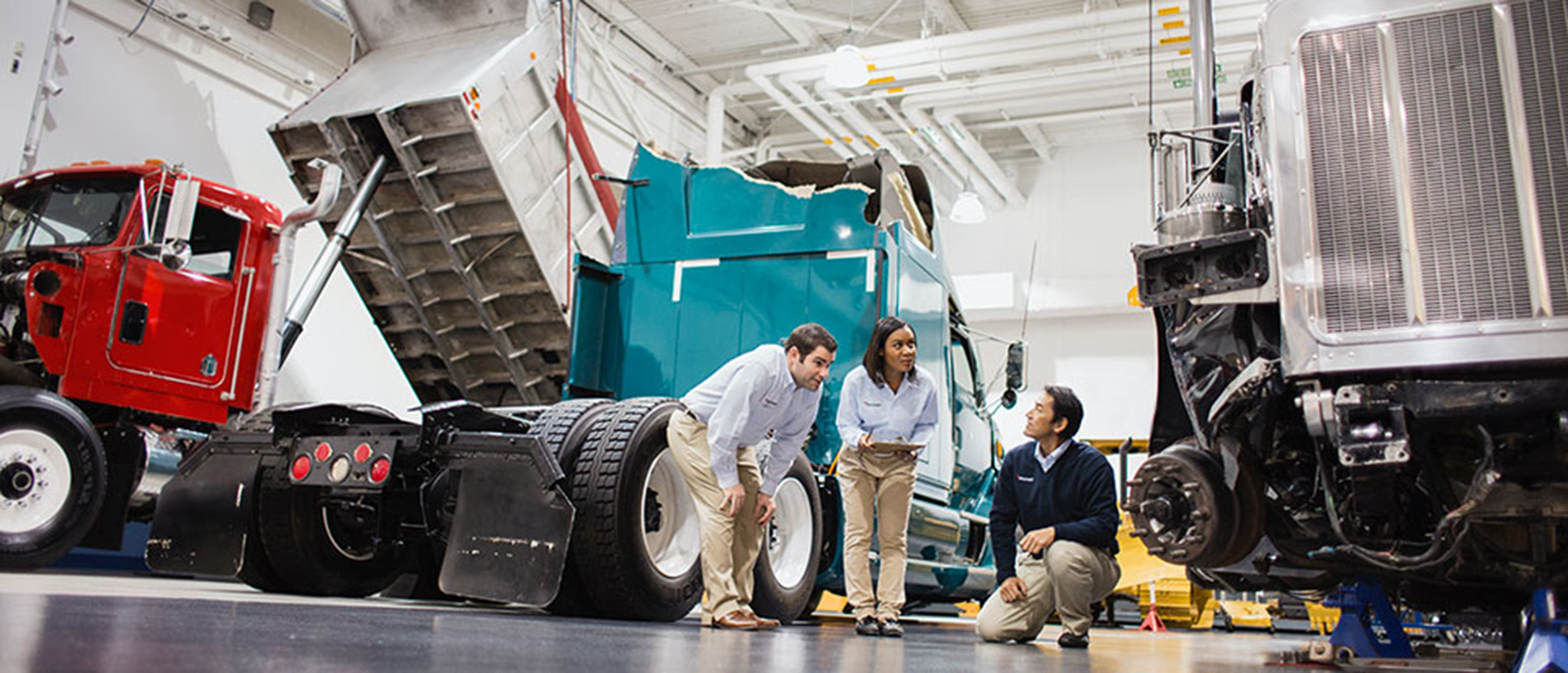 Female manager holding a digital tablet and talking with two male colleagues in large warehouse of trucks.