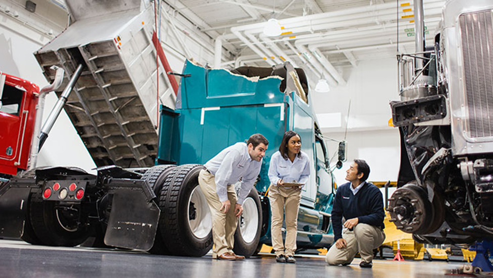 Female manager holding a digital tablet and talking with two male colleagues in large warehouse of trucks.