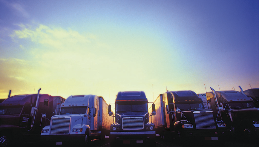 fleet of large semi trucks lined up in a row, with the sun setting behind them