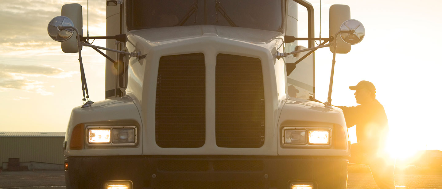 Front of a white truck, headlights on, with the silhouette of the driver in front of an evening sunset.