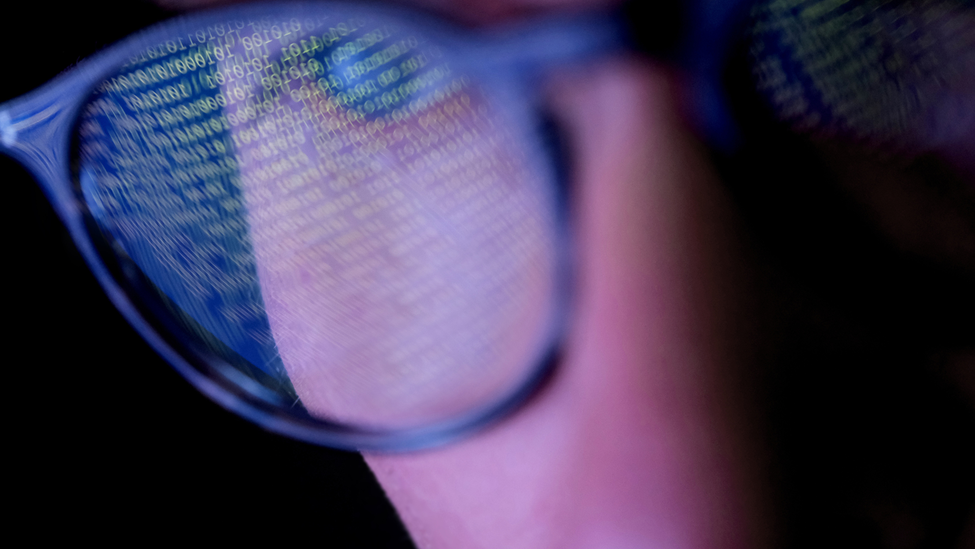 A person with glasses in a dark room looking at computer screen that's displaying codes or programming, with reflections on the glasses