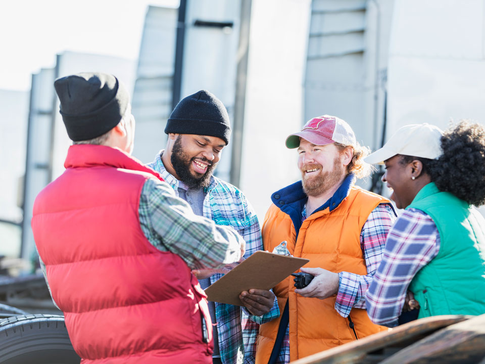 Group of drivers standing in front of a fleet of semitrucks, laughing, holding a clipboard.