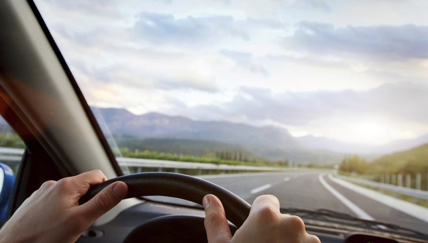 Closeup of two hands on car steering wheel driving safely down a road.