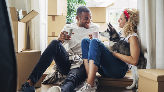 A man and woman sitting on floor with dog surrounding by boxes.