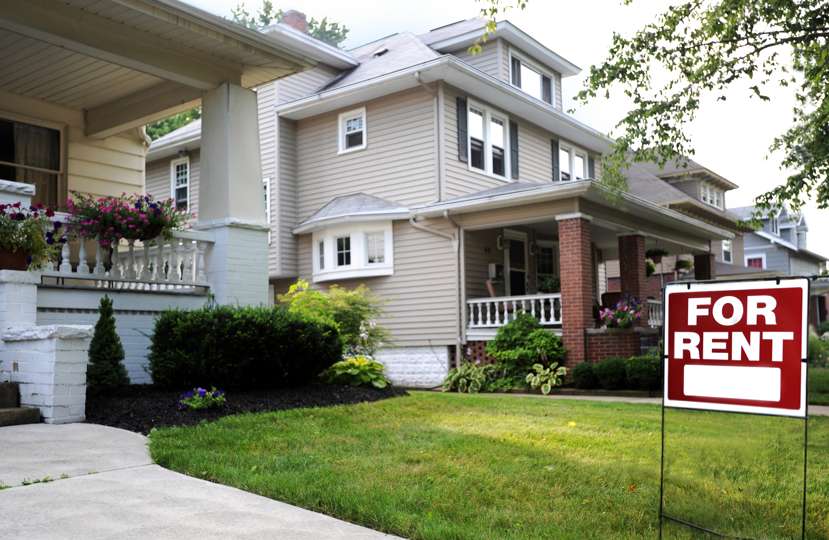 A "For Rent" sign posted on the front lawn of a bungalow style house.