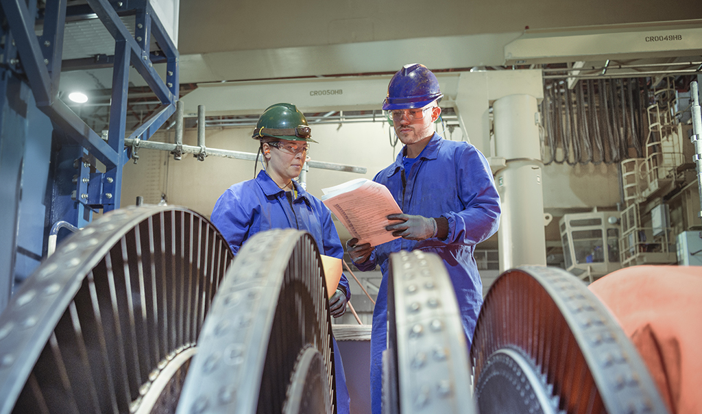 Two workers in safety gears having a discussion and checking a machine with gears.