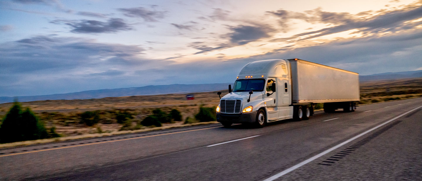 Long haul semitruck with headlights on driving on the highway to make deliveries under an evening sky.
