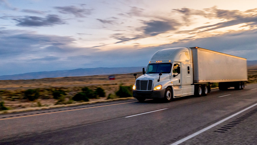 Long haul semitruck with headlights on driving on the highway to make deliveries under an evening sky.