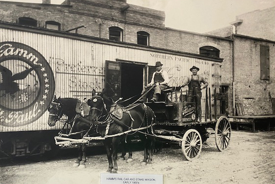 Male sitting and holding the reins for two dark horses while another male stands behind him on a stake wagon. They are standing in front of a Hamm’s rail car in the early 1900s.