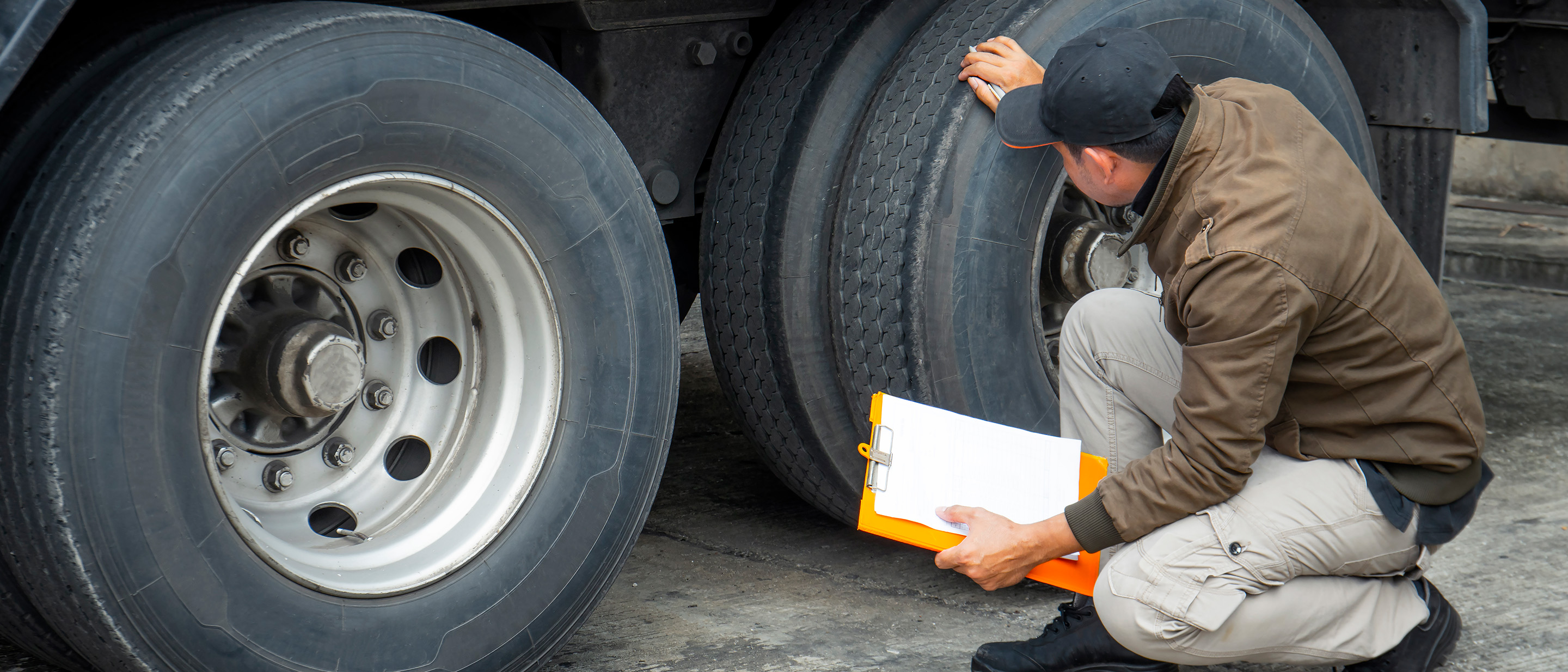 Male truck driver holding a clipboard stooping down for a safety check on truck tires.